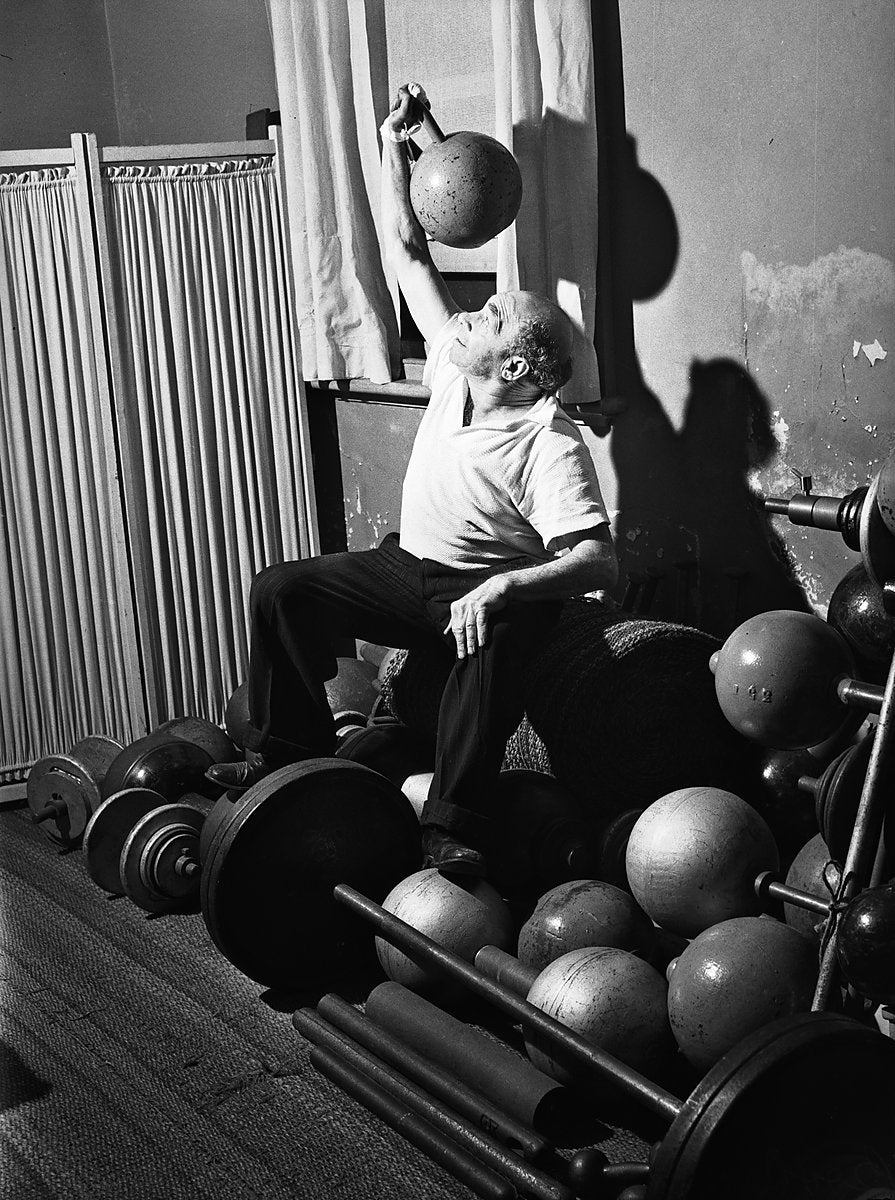 Man pressing a globe dumbbell overhead among barbells and weights — photograph, from Marrickville, 1949