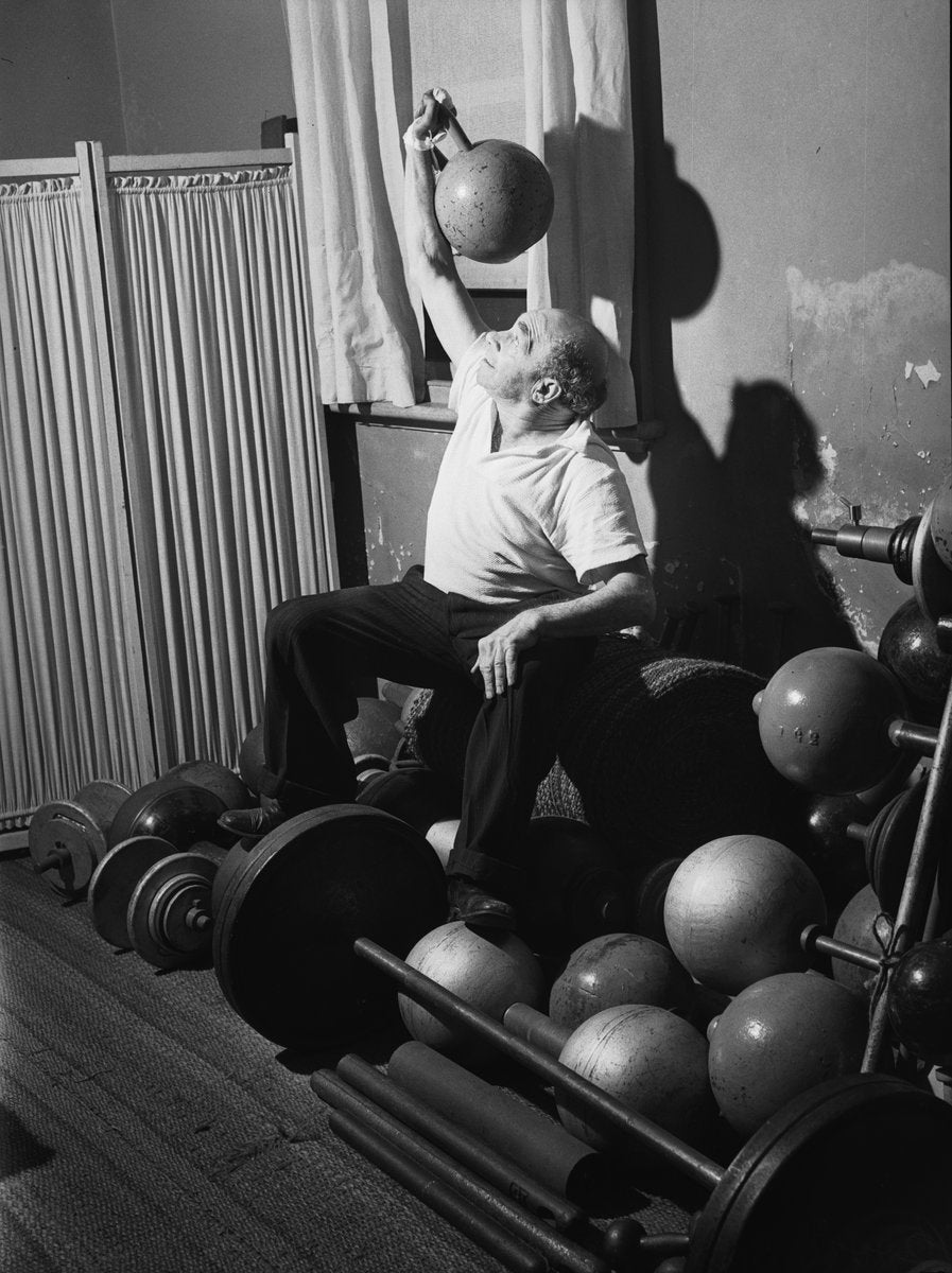 Man pressing a globe dumbbell overhead among barbells and weights — photograph, from Marrickville, circa 1949