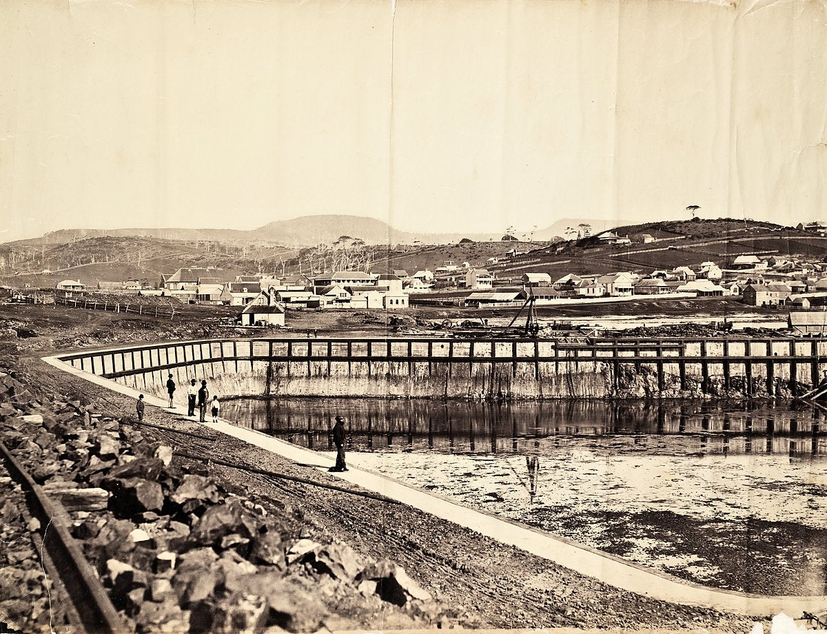 Water-filled basin under construction below hillside houses — photograph, 1858–1900