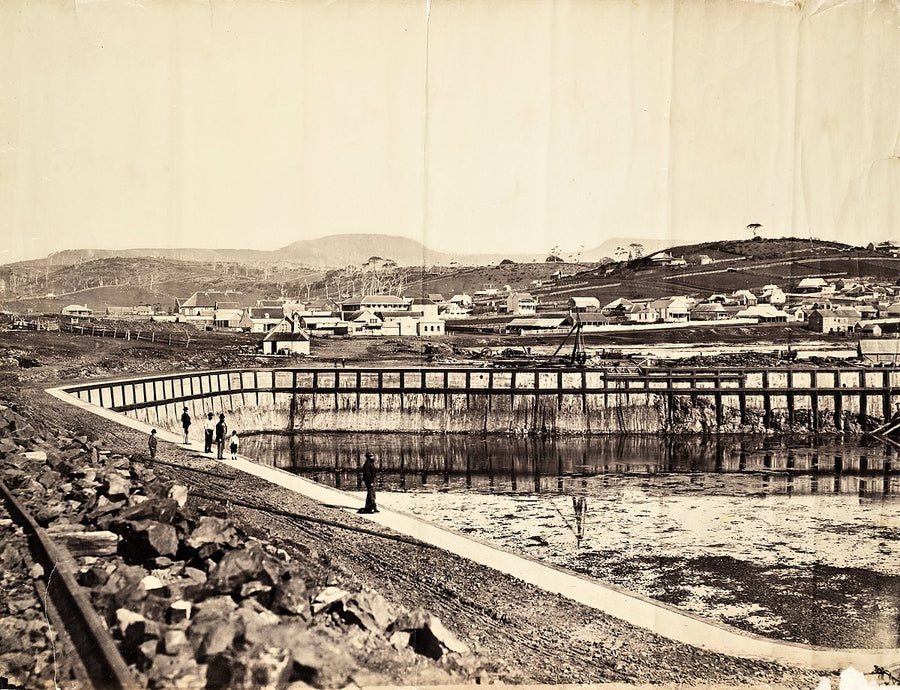Water-filled basin under construction below hillside houses — photograph, 1858–1900