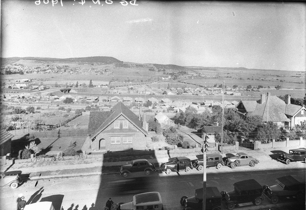 Elevated view across a township to low hills, motor cars parked along the road — photograph, from null, circa 1930